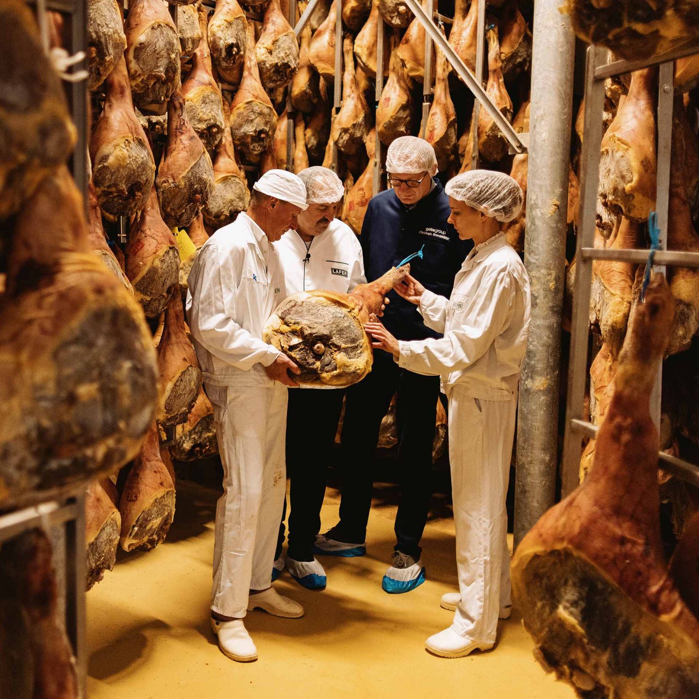 Three workers in protective clothing inspect a large cured ham in a storage room filled with hanging hams.