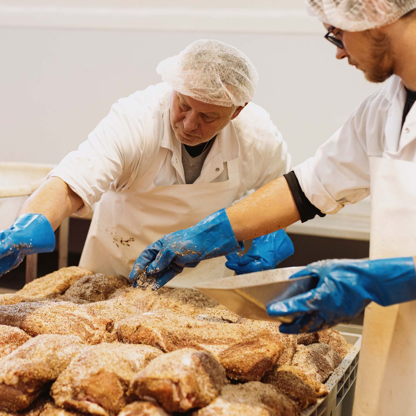 Two workers in hairnets and gloves apply seasoning to rows of meat in a food processing facility.
