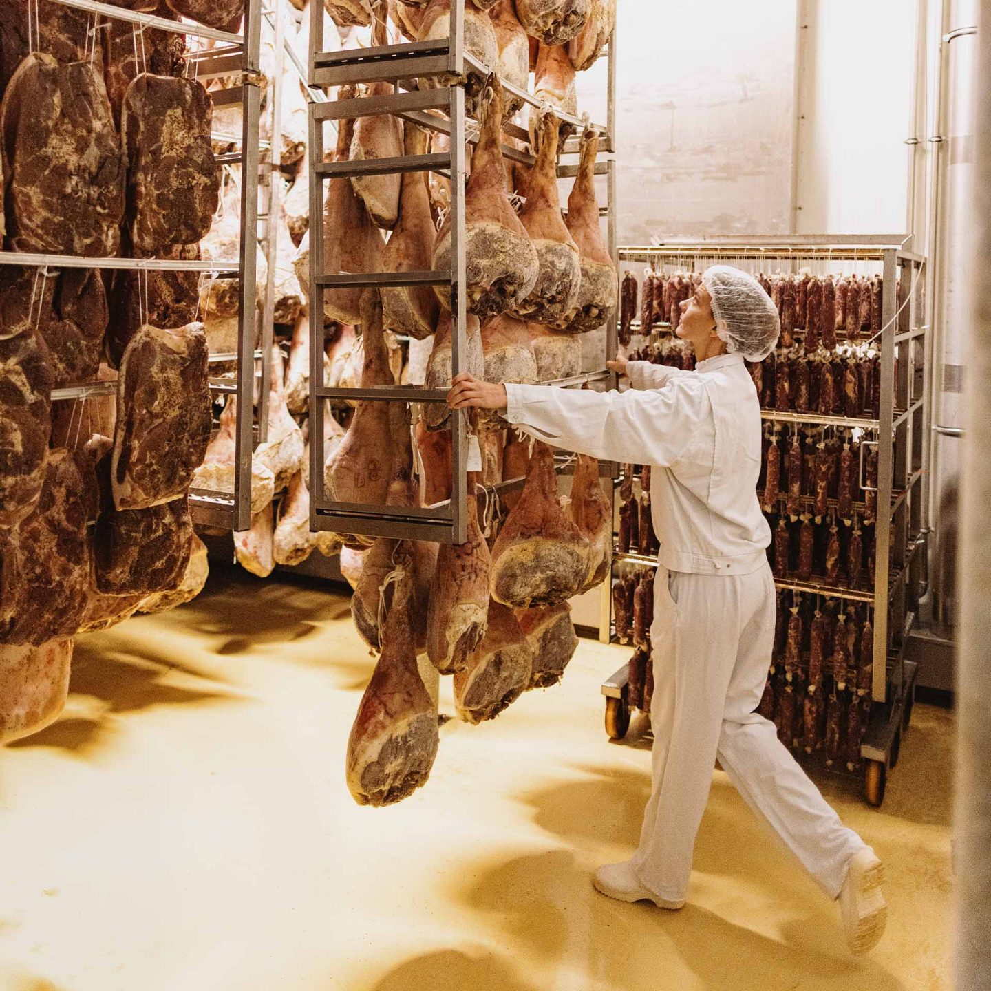 Worker in protective clothing handles hanging cured hams in a meat curing facility with racks of sausages.