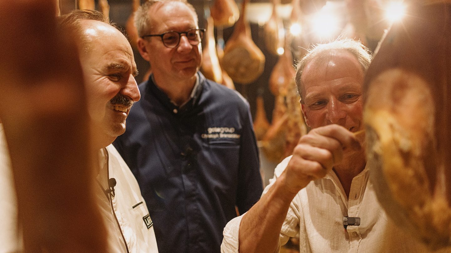 Three men inspect hanging cured hams in a traditional meat curing cellar.