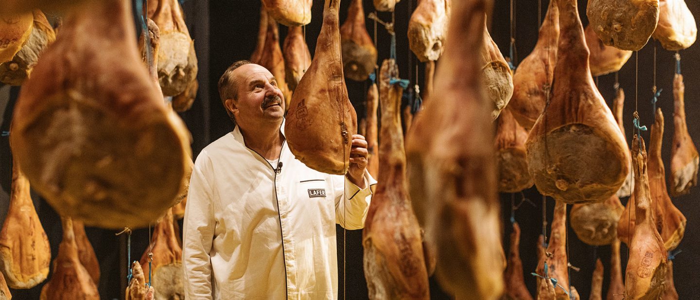 A man inspects hanging cured hams in a traditional meat curing room with dim lighting.
