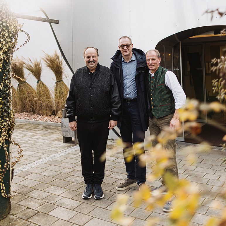 Three men stand outdoors on a paved area with bundled wheat plants and a building entrance in the background.