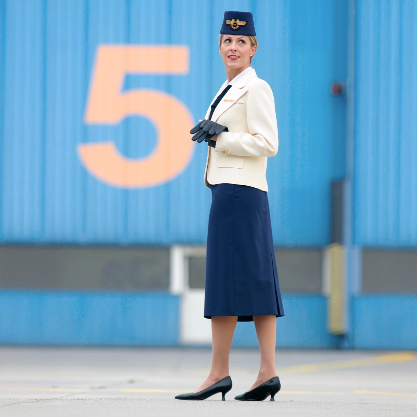 Flight attendant in previous Lufthansa uniform with light blazer, dark skirt, and badge hat.