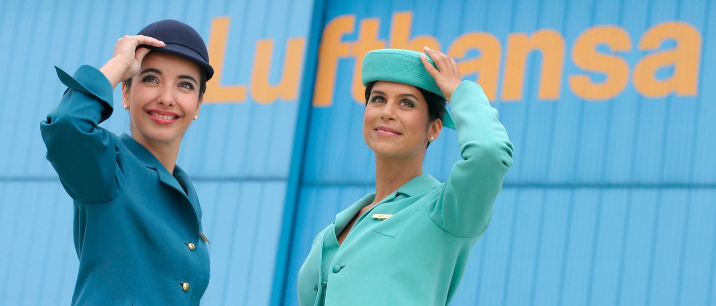 Two Lufthansa flight attendants in teal and navy uniforms with hats stand in front of a Lufthansa sign.