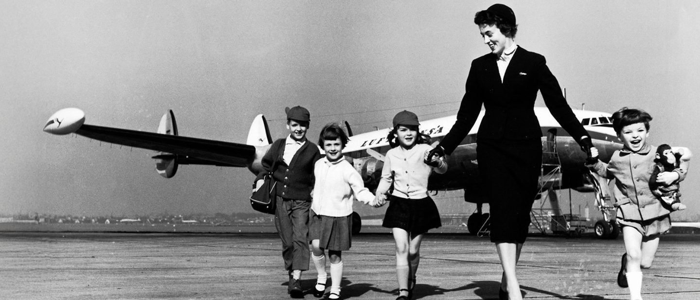 A flight attendant walks with children across the tarmac in front of a Lufthansa aircraft.
