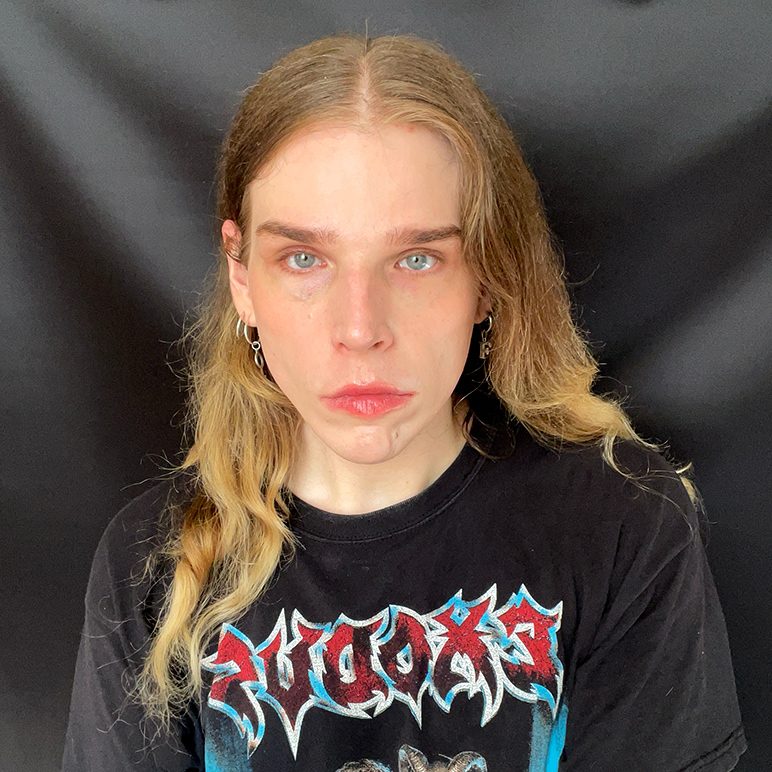 A young person with blond hair wearing a black band T-shirt, in front of a dark background