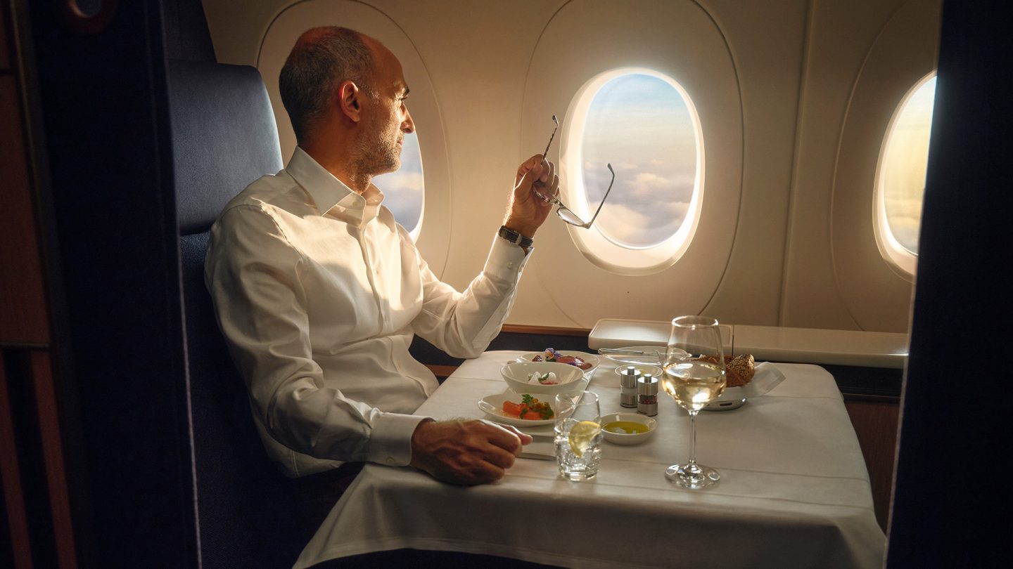 Man in first class sits by the window eating, drink on the table.