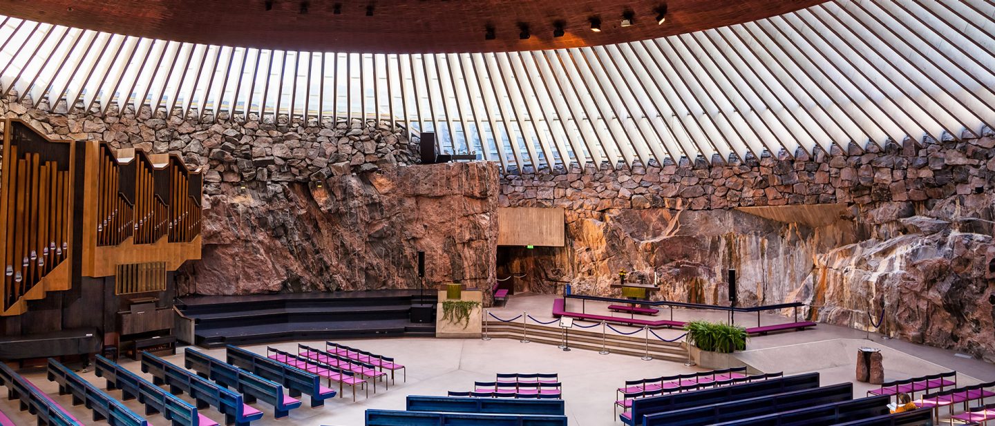 Interior view of a modern church with stone walls and wooden organ pipes under a radial skylight.
