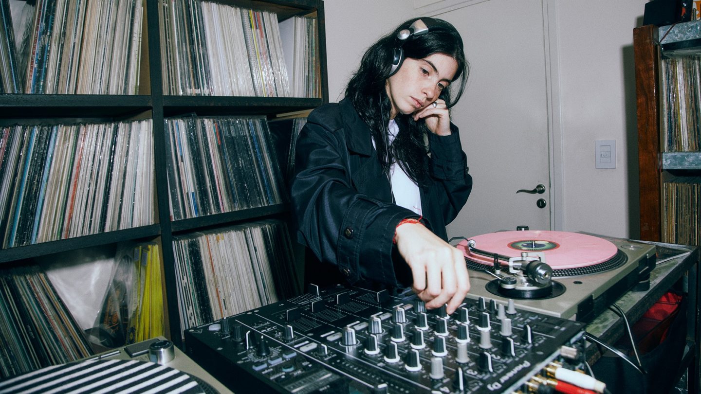DJ Franzizca plays records and works intently at a mixing desk, surrounded by a large shelf full of vinyl records.