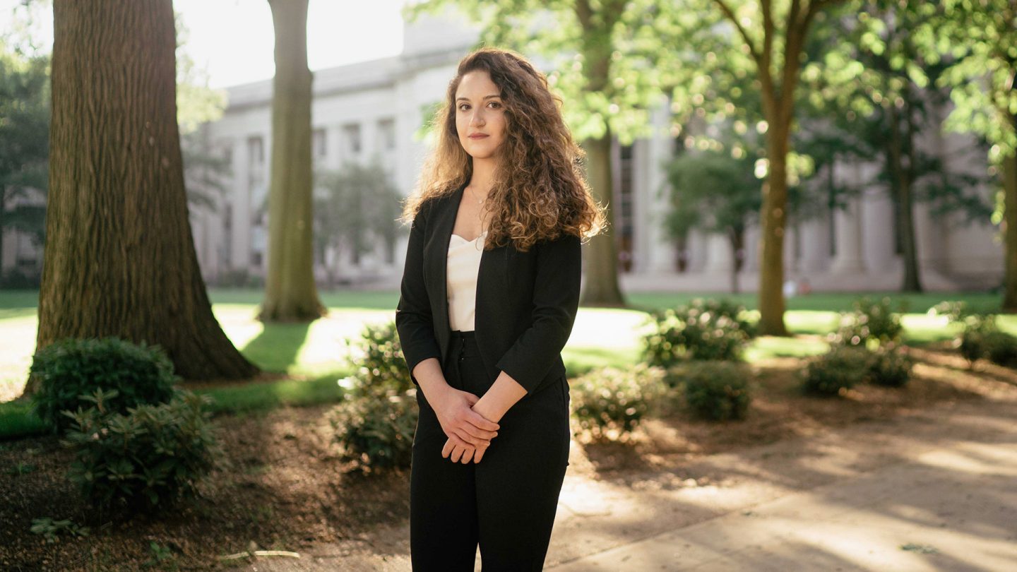 Portrait of Maya Nasr, with curly hair, wearing a black pantsuit, standing in a sunny park in front of a neoclassical building