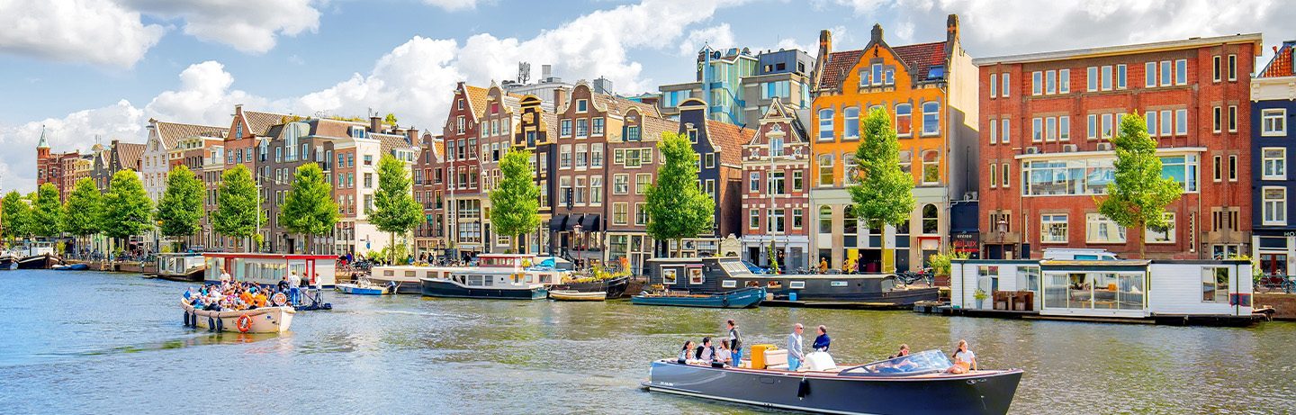Several boats are navigating a canal in Amsterdam. Colorful historic gabled houses stand on the banks.