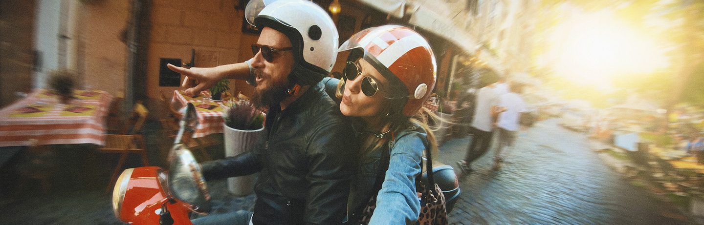 A couple on a scooter rides through a sunny old town street with cafés and evening light
