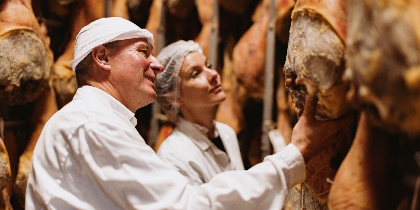 Two people in white coats and hairnets inspect hanging cured ham in a meat aging facility.