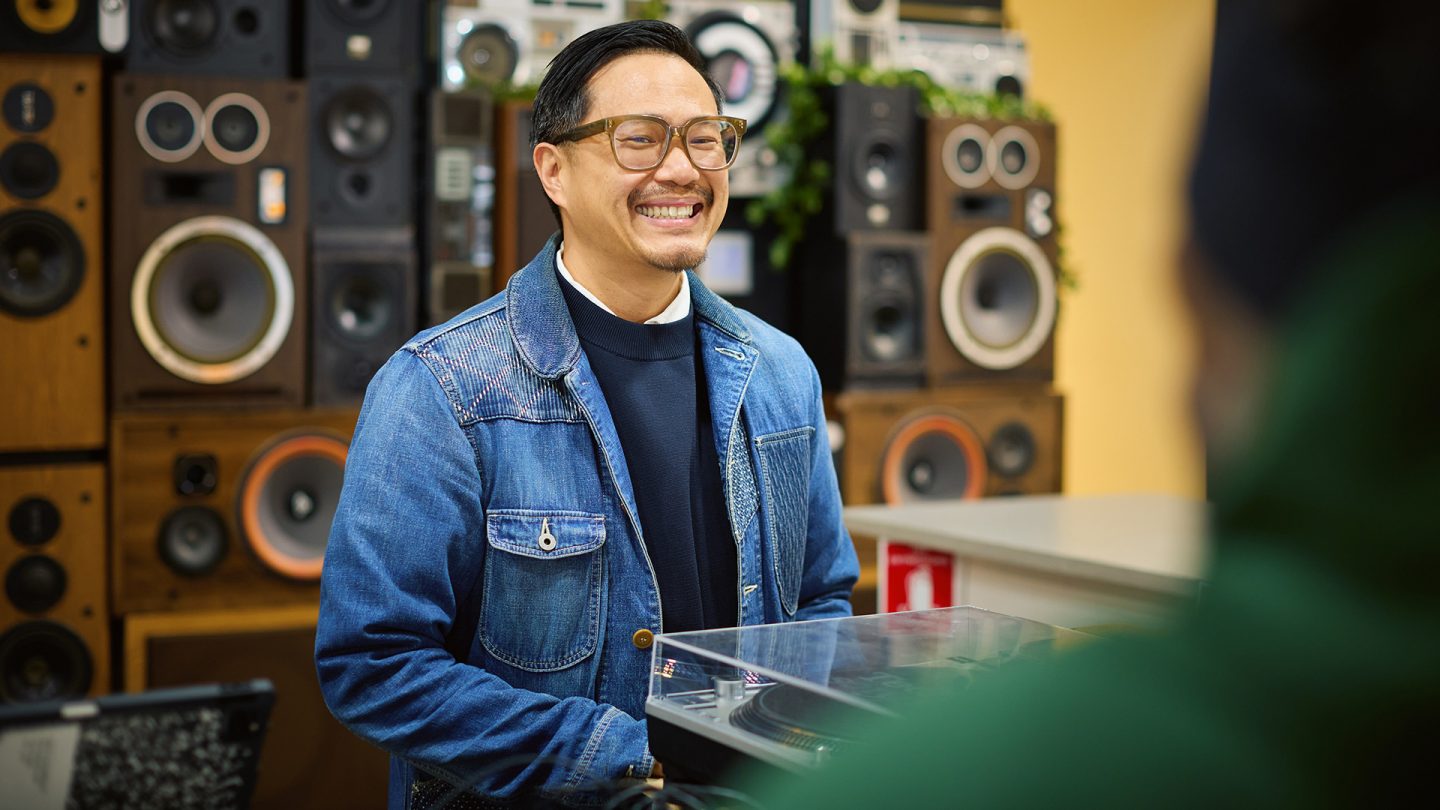 Oliver Mak, wearing glasses and a denim jacket, stands in front of a wall of speakers and smiles