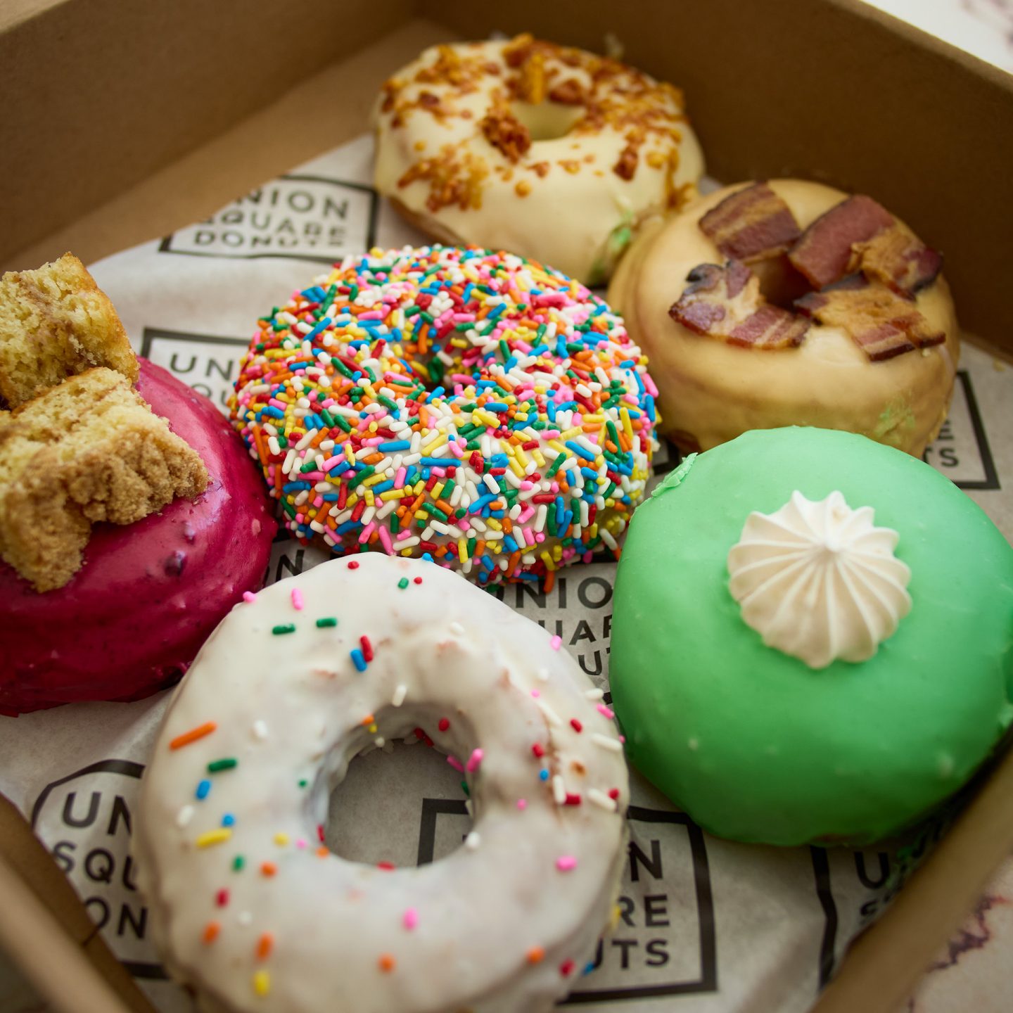 A box of six donuts, including varieties with sprinkles, bacon, green icing, and berry glaze